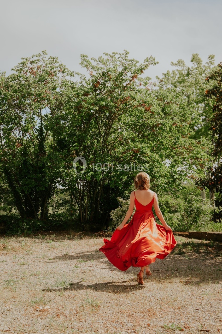 Une femme en robe rouge marche dans un jardin, entourée d'arbres et de verdure sous un ciel clair.