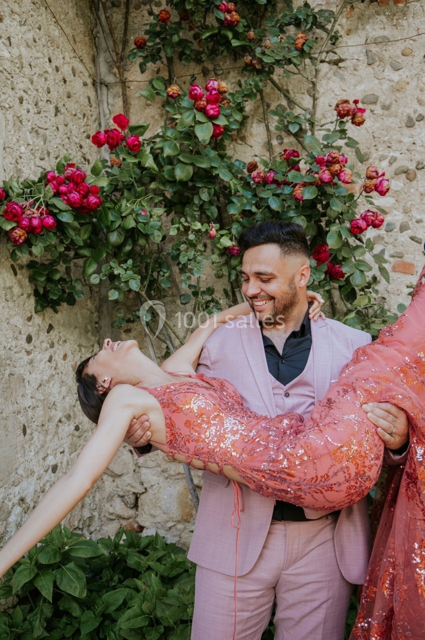 Un homme en costume rose porte une femme en robe scintillante devant un mur en pierre orné de rosiers fleuris.