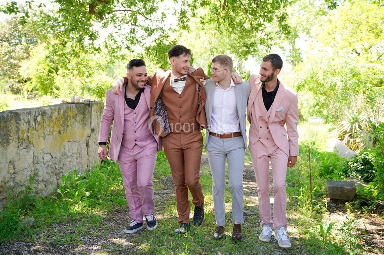 Quatre hommes en tenue élégante marchent ensemble sur un chemin bordé de verdure, souriants et détendus.