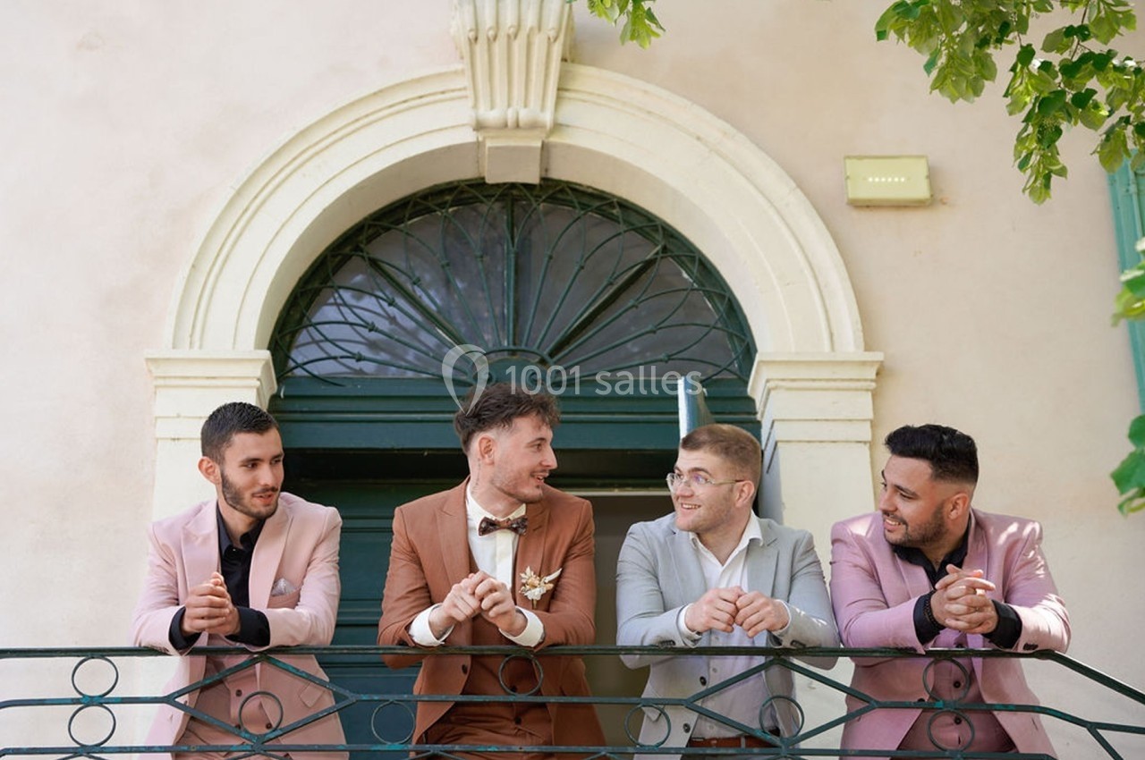 Quatre hommes en costumes élégants discutent et sourient sur un balcon devant une porte voûtée.