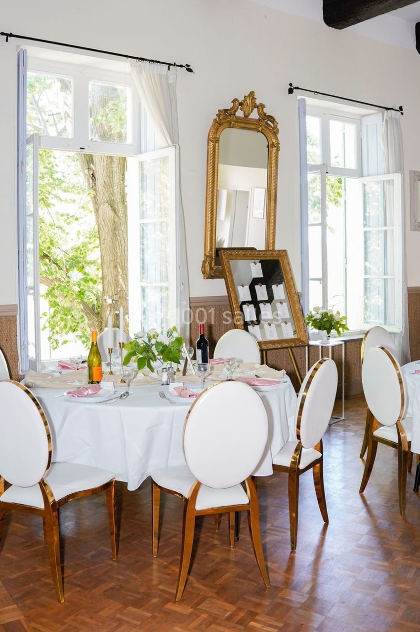 Salle lumineuse avec tables rondes dressées, chaises blanches, grand miroir doré et fenêtres ouvertes sur un jardin.
