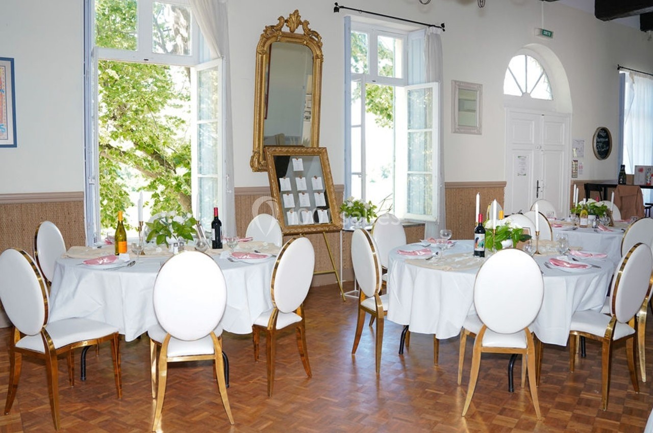 Salle de réception lumineuse avec tables rondes dressées, chaises blanches et grandes fenêtres donnant sur un jardin.
