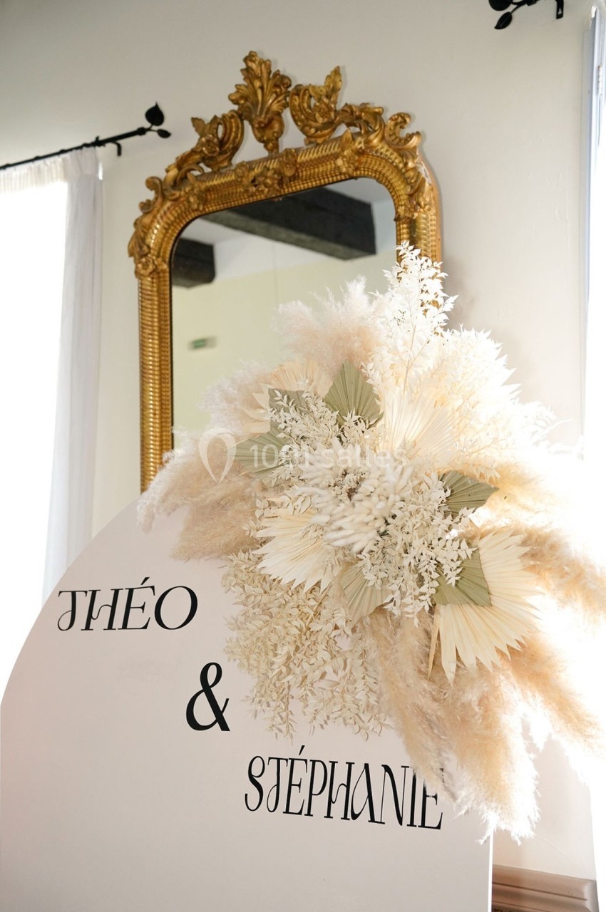 Décoration florale blanche et beige devant un miroir doré, avec les prénoms ’Théo & Stéphanie’ inscrits sur un support.