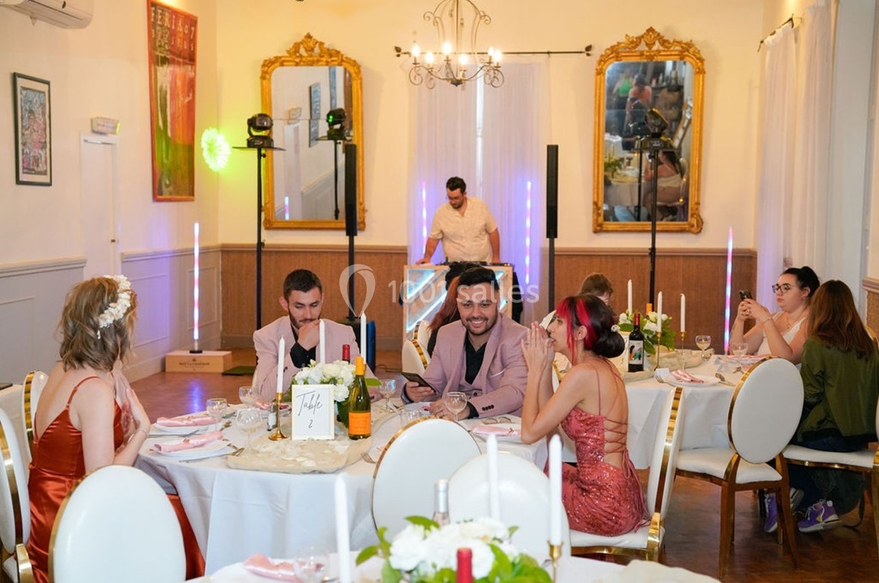 Des convives assis autour de tables décorées dans une salle élégante, avec un DJ en arrière-plan.