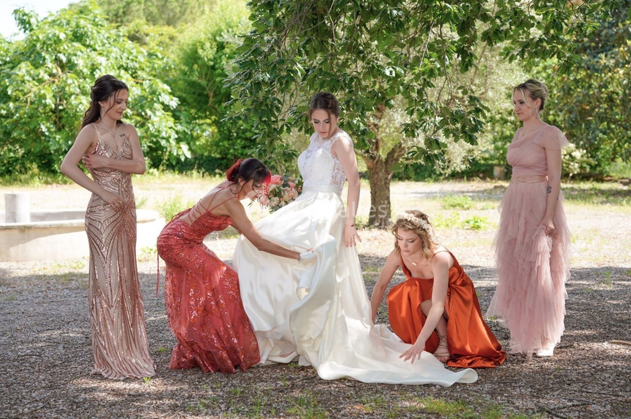 Une mariée en robe blanche entourée de quatre femmes ajustant sa tenue dans un jardin verdoyant.