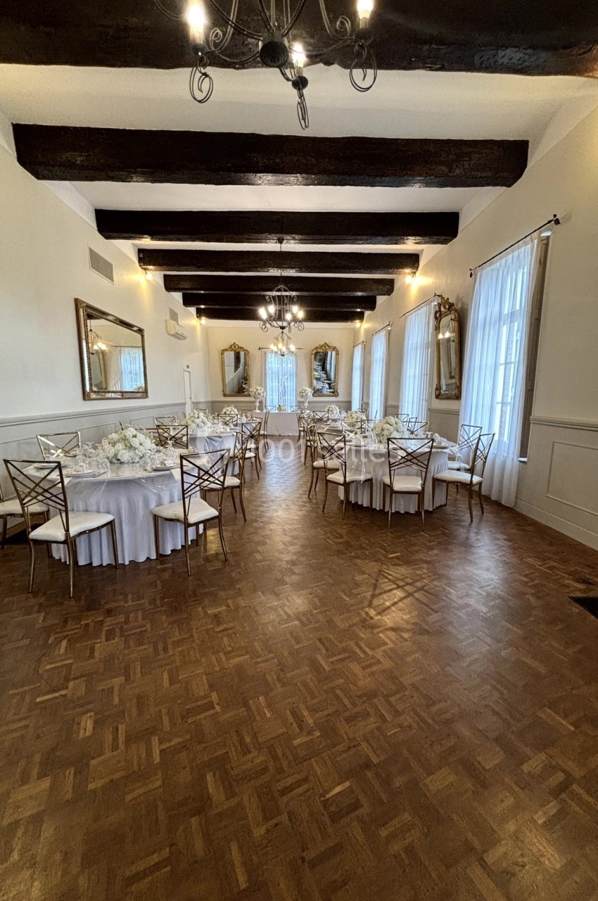 Salle de réception élégante avec parquet, tables rondes dressées, chaises dorées et poutres apparentes au plafond.