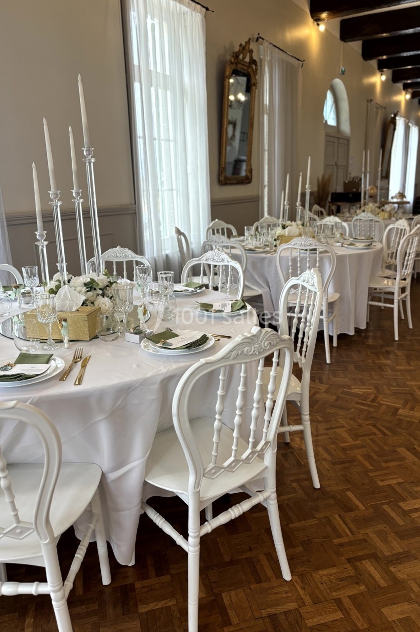 Salle de réception élégante avec tables rondes dressées, chaises blanches et décorations florales raffinées.