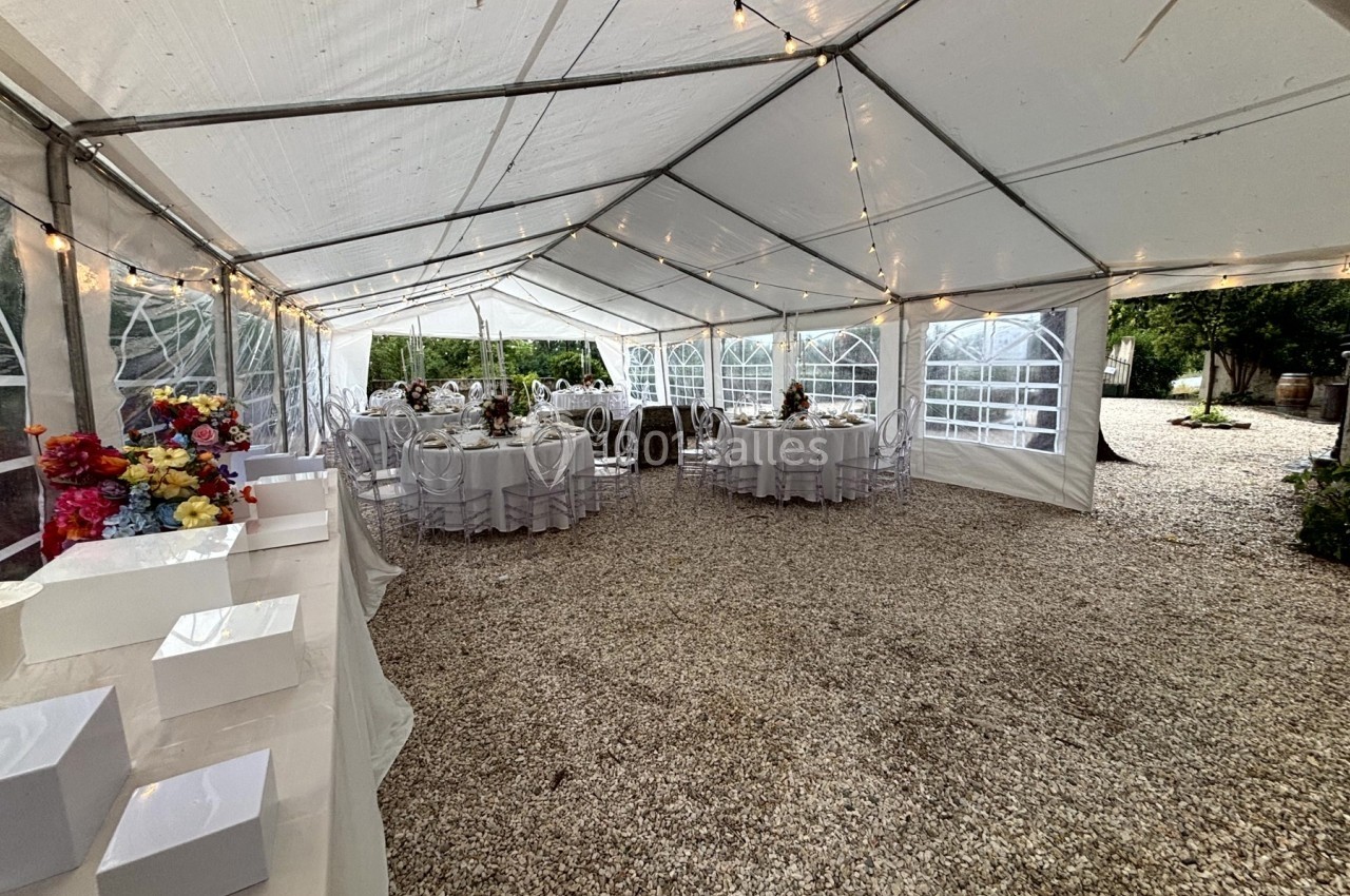 Intérieur d'une tente blanche avec des tables rondes dressées pour un événement, éclairée par des guirlandes lumineuses.