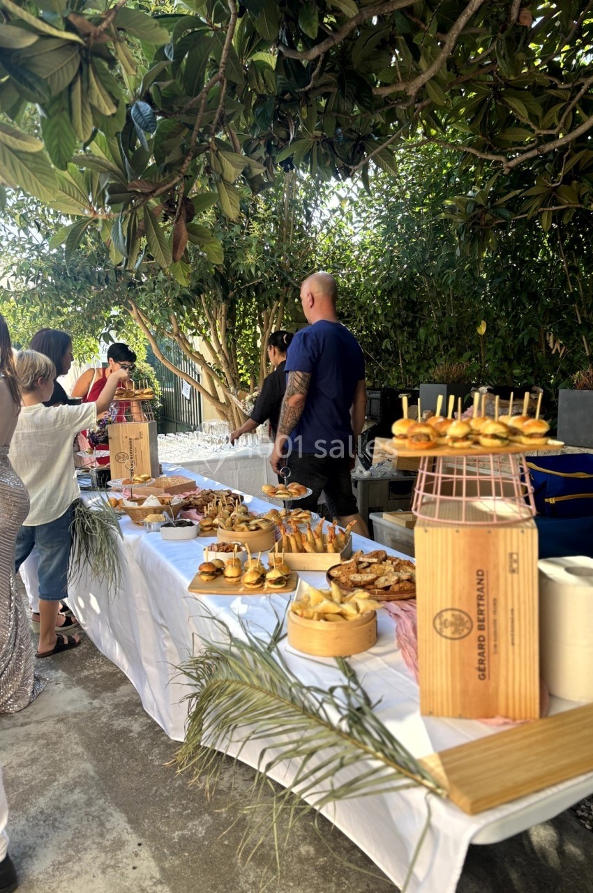 Table de buffet en extérieur avec divers plats et amuse-bouches, entourée de convives dans un jardin arboré.