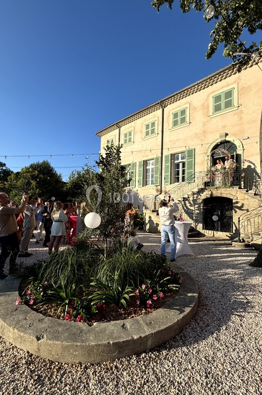 Jardin avec parterre fleuri devant une grande maison en pierre, des personnes discutent et prennent des photos.