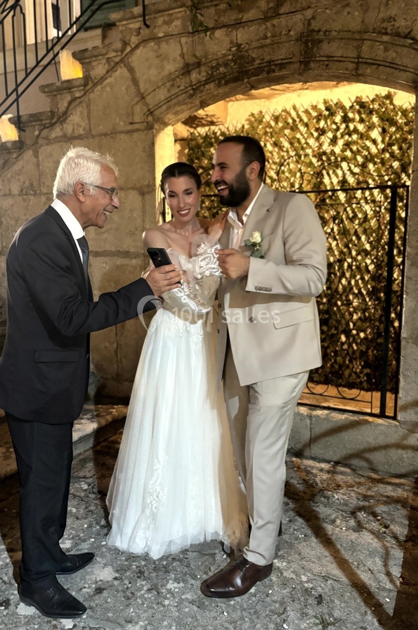 Un couple en tenue de mariage regarde un téléphone avec un homme âgé, debout devant un mur en pierre éclairé.