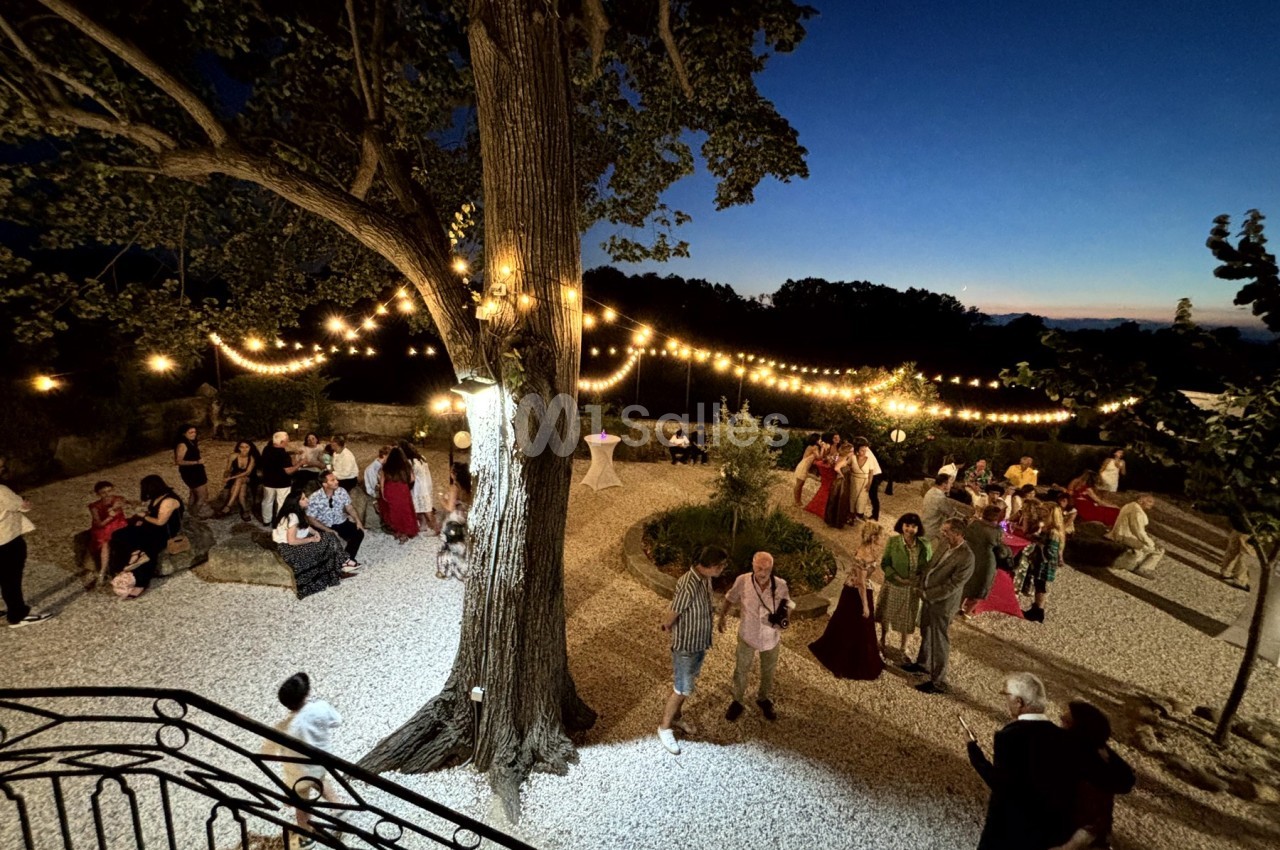 Personnes rassemblées en soirée dans un jardin éclairé par des guirlandes lumineuses, sous un grand arbre.