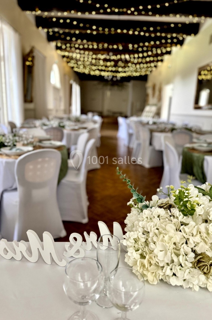 Salle de réception décorée avec des guirlandes lumineuses, des tables dressées et un bouquet de fleurs blanches au premier…