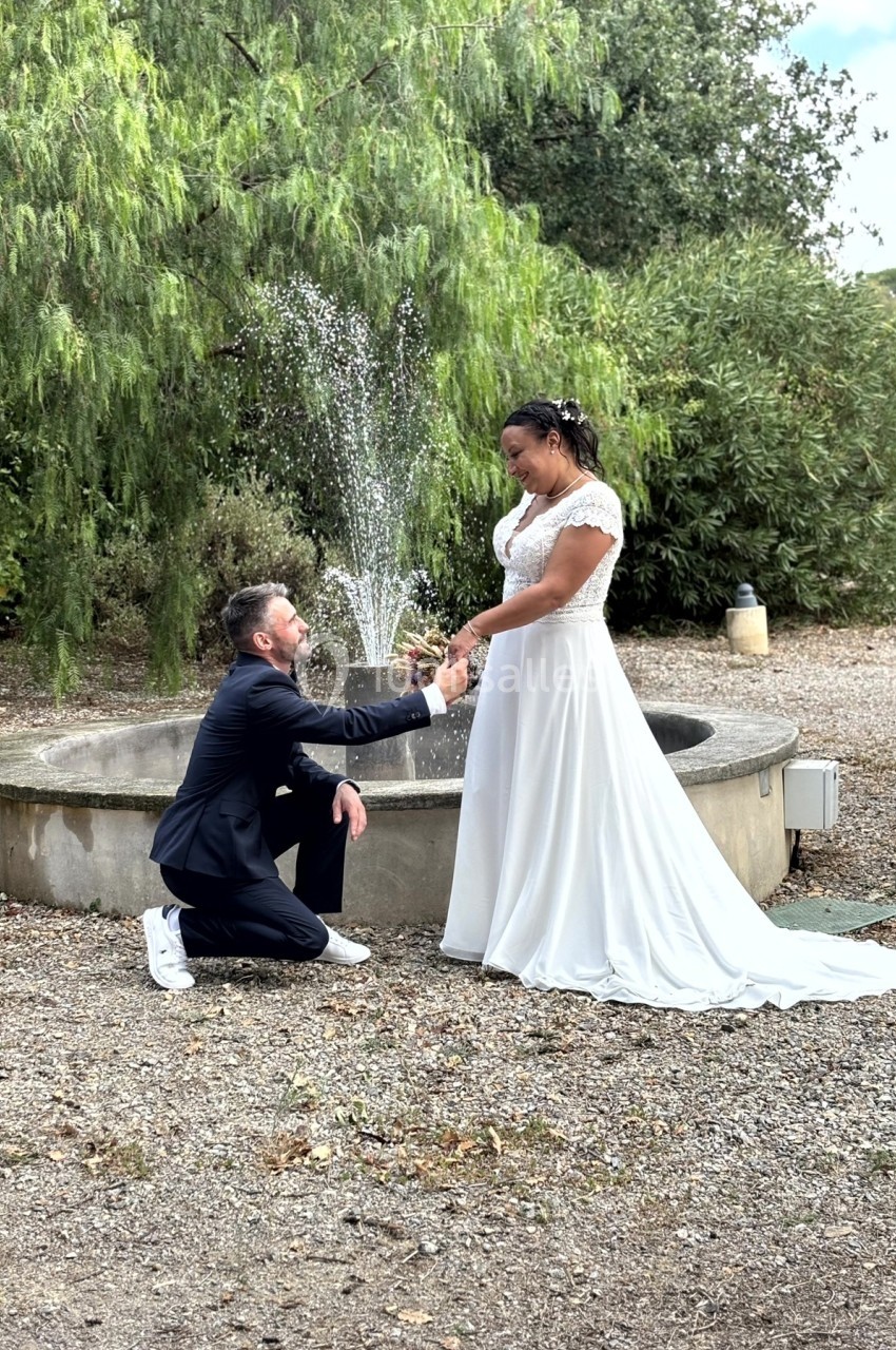 Un homme en costume propose à une femme en robe blanche devant une fontaine dans un jardin.