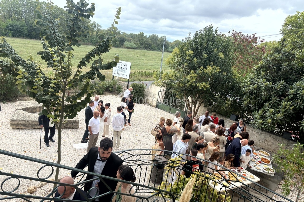 Groupe de personnes rassemblées dans une cour extérieure autour de tables de buffet, avec un paysage verdoyant en arrière…