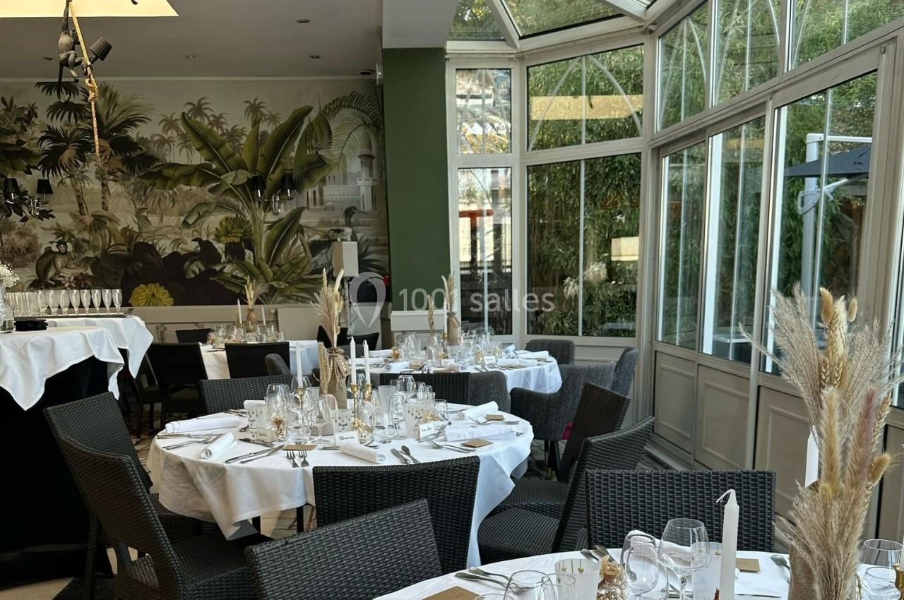 Salle de réception lumineuse avec tables rondes dressées, décor végétal et grandes baies vitrées donnant sur l'extérieur.