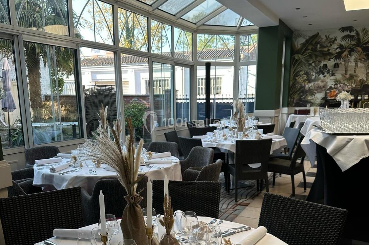Salle lumineuse avec verrière, tables dressées avec nappes blanches, décorations florales et chaises en osier.