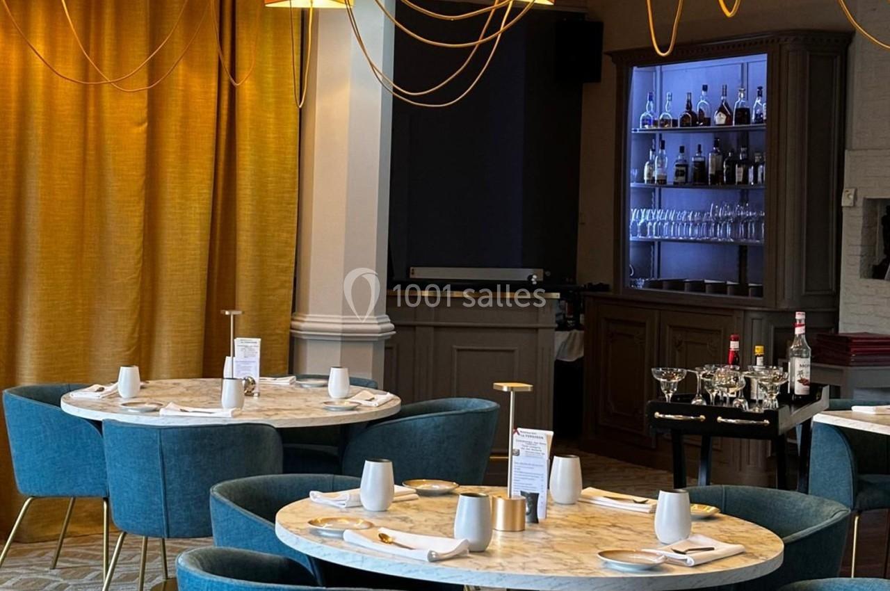 Salle de restaurant élégante avec tables rondes en marbre, chaises bleues et un bar illuminé en arrière-plan.