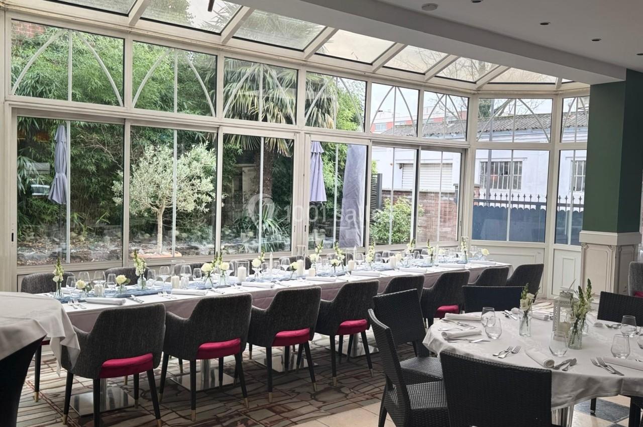 Salle lumineuse avec de grandes baies vitrées, tables dressées pour un repas, vue sur un jardin extérieur.