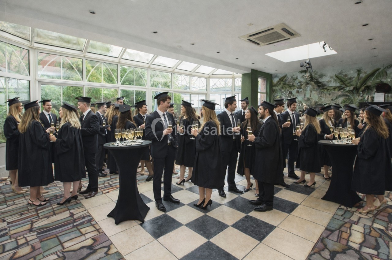 Des étudiants en tenue de remise de diplômes discutent et trinquent dans une salle lumineuse avec des tables hautes.