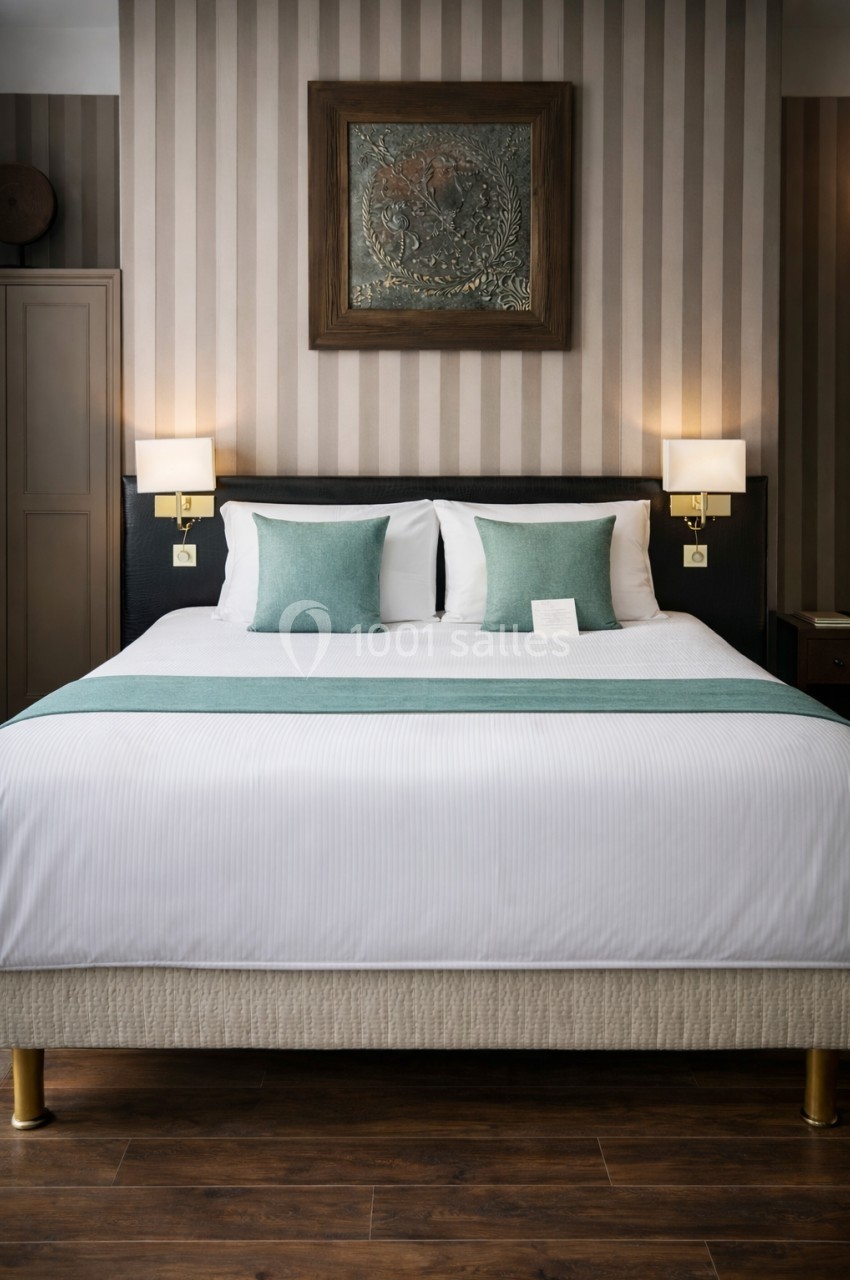 Lit double avec draps blancs, coussins verts, deux lampes murales et un tableau encadré sur un mur à rayures.