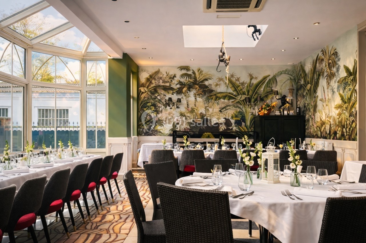 Salle de réception lumineuse avec tables dressées, chaises en osier, fresque murale tropicale et verrière.