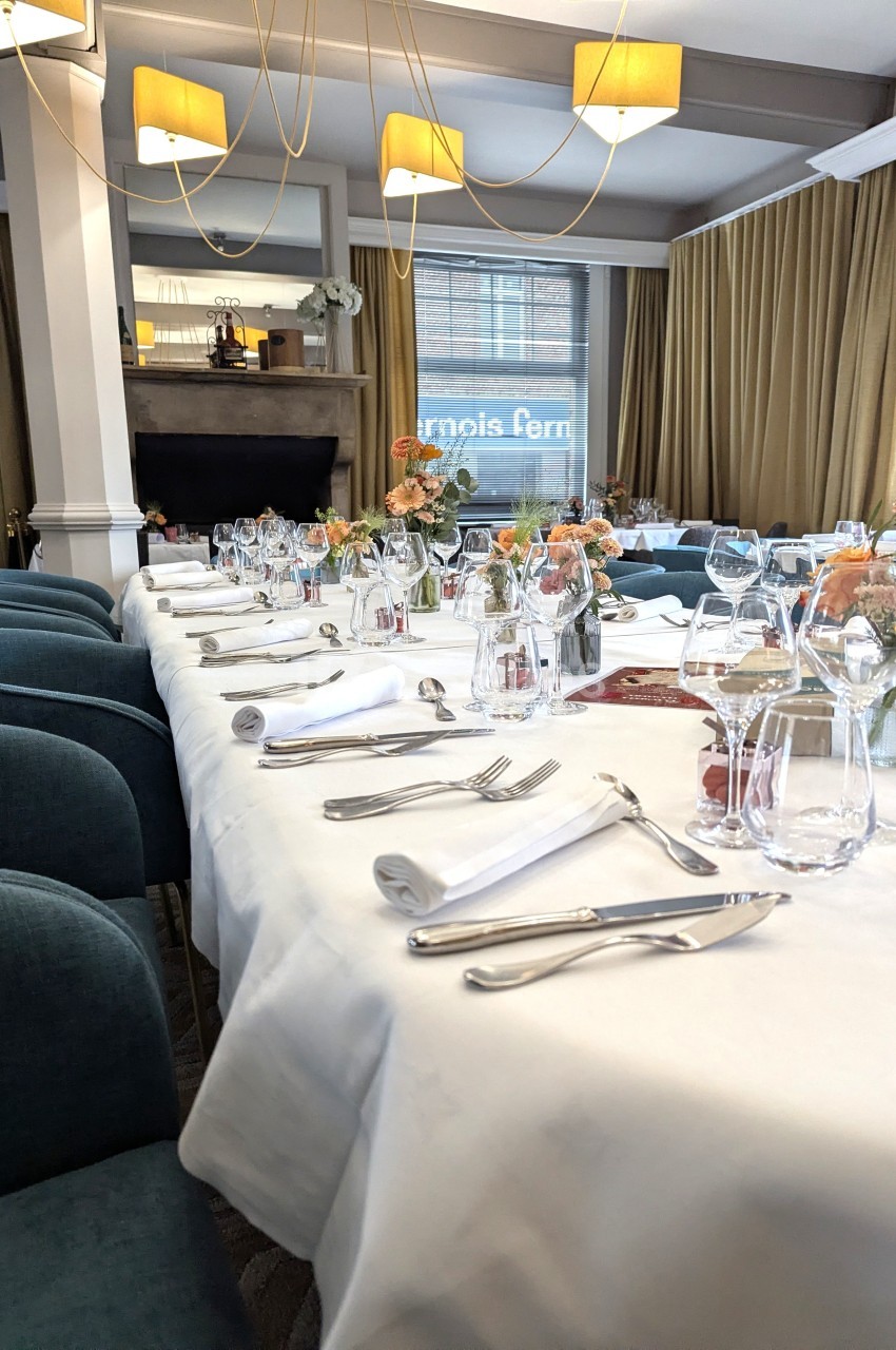 Table dressée avec nappes blanches, couverts, verres et décorations florales dans une salle lumineuse avec grandes fenêtres.