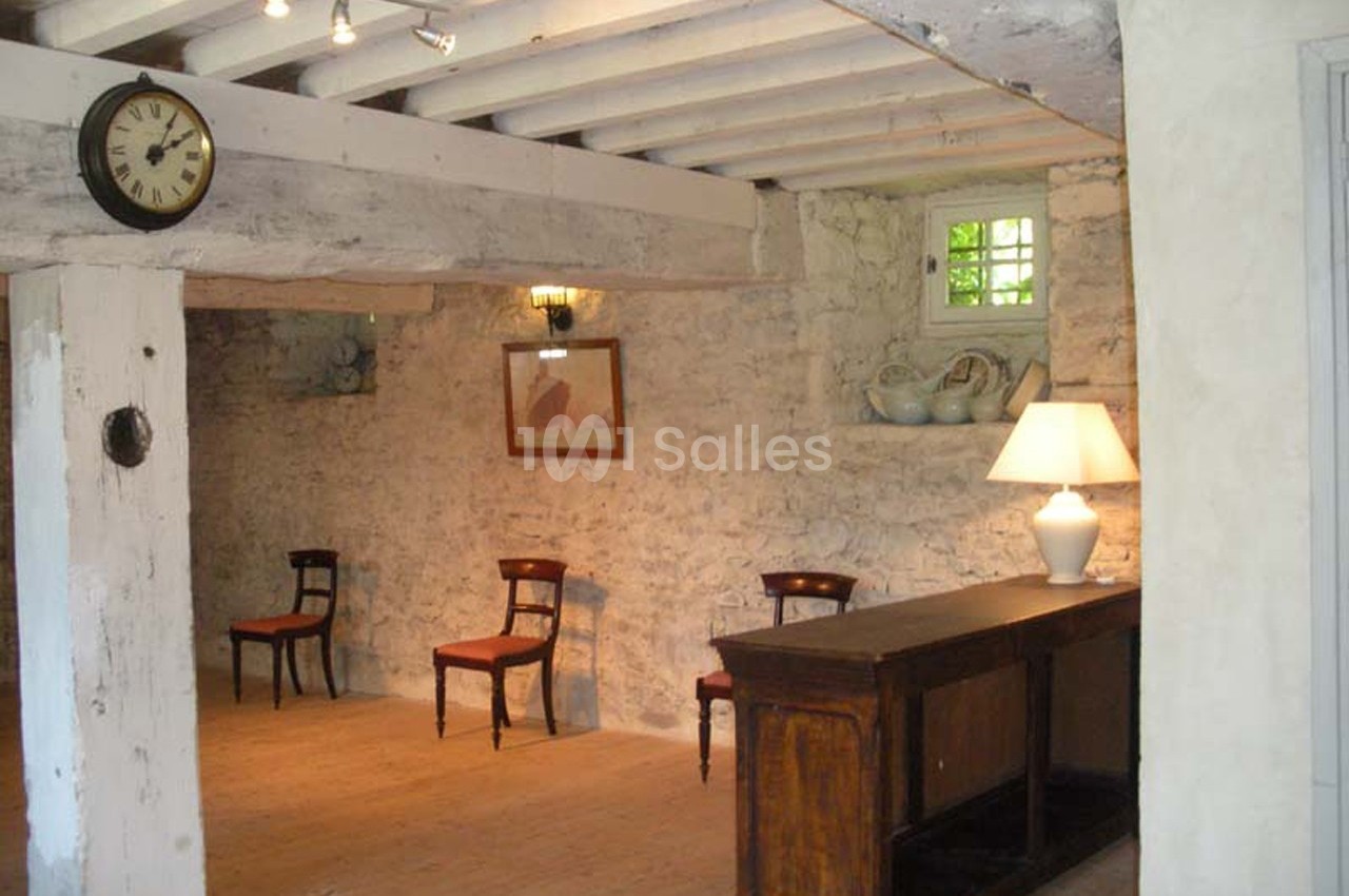 Salle rustique avec murs en pierre, poutres apparentes, mobilier ancien, trois chaises en bois et une lampe sur un comptoir.