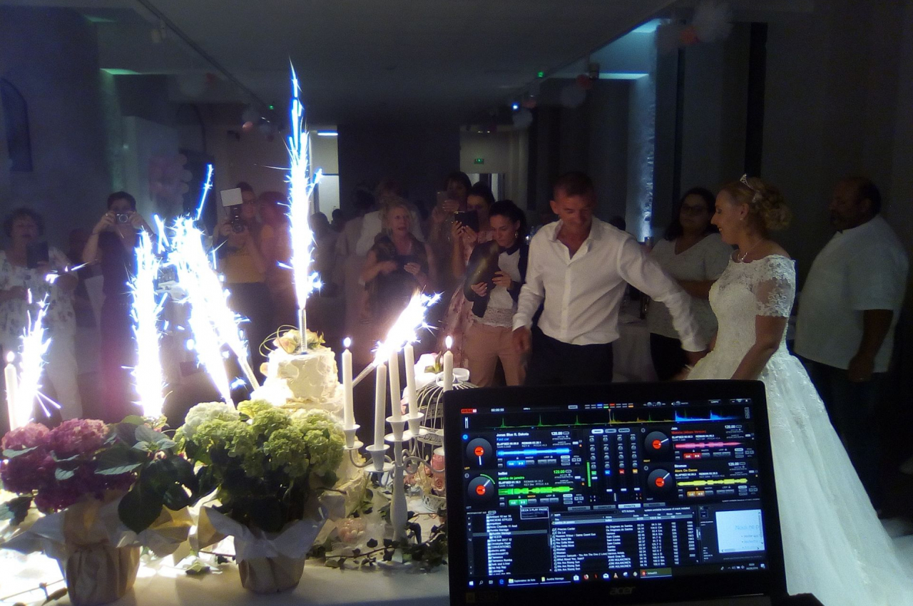 Un couple de mariés se tient devant un gâteau entouré de bougies et de fontaines lumineuses, avec des invités en arrière…