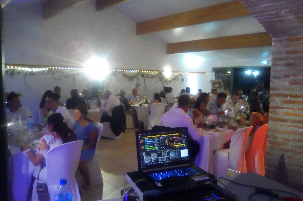 Salle de réception éclairée avec des invités assis à des tables décorées, vue depuis une table de DJ avec un ordinateur.