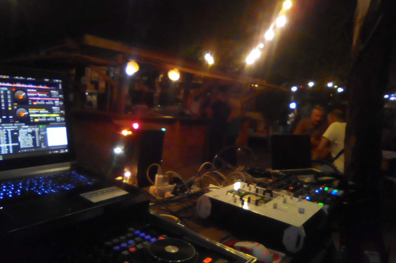 Poste de DJ avec ordinateurs et tables de mixage, éclairé par des guirlandes lumineuses dans un espace extérieur nocturne.