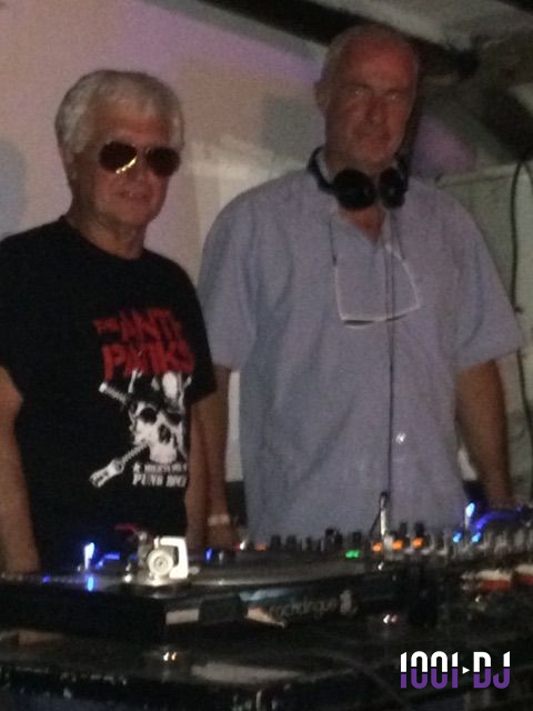 Deux hommes debout derrière une table de mixage avec platines, l'un portant des lunettes de soleil.