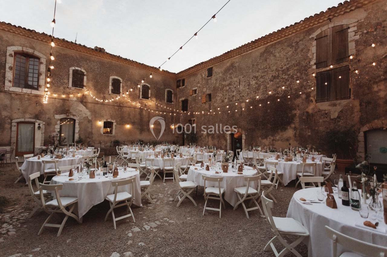 Cour intérieure rustique avec tables rondes dressées pour un événement, éclairée par des guirlandes lumineuses suspendues.