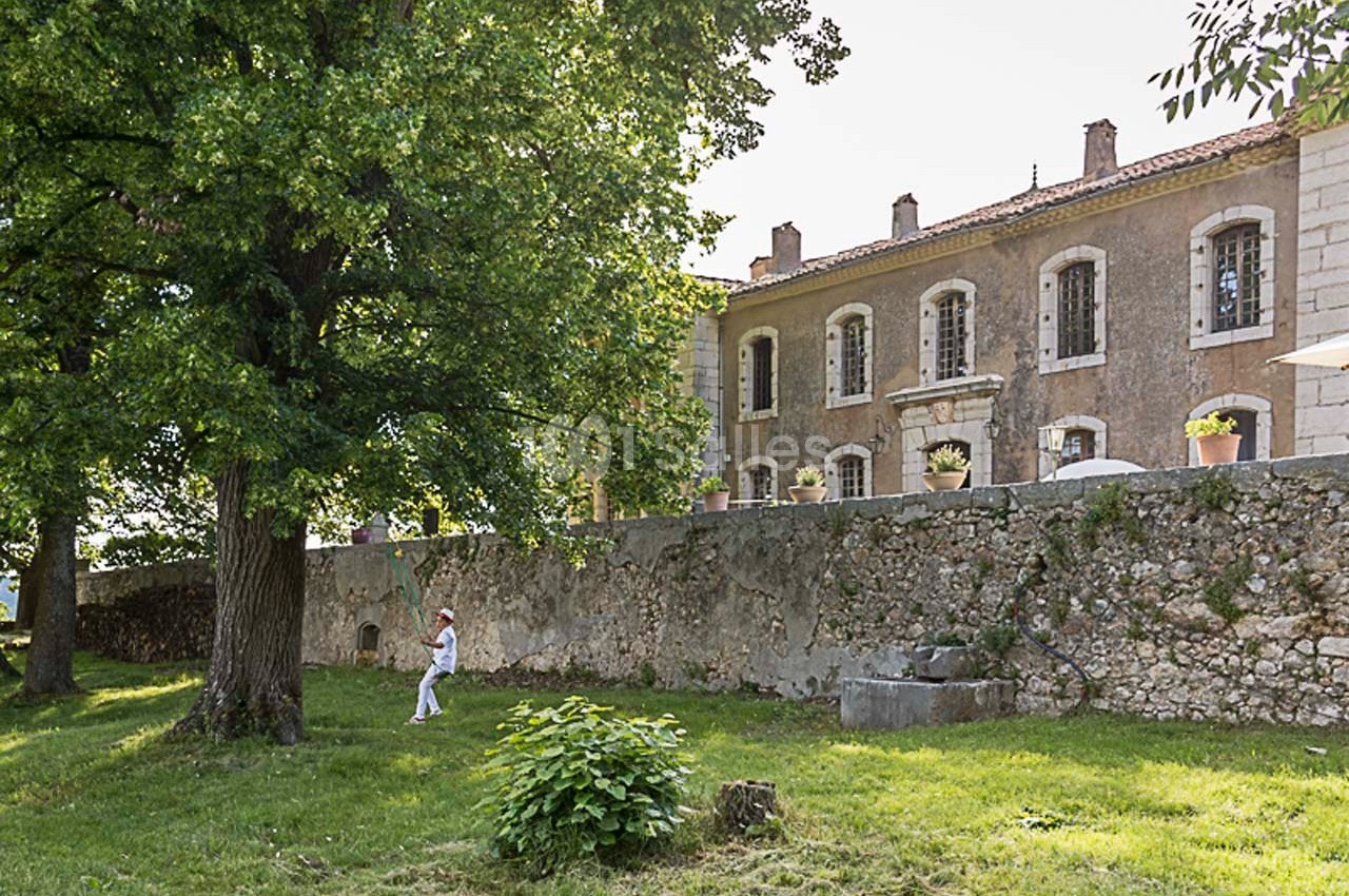 Façade d'un bâtiment en pierre avec des volets, vue depuis un jardin arboré sous un ciel clair.