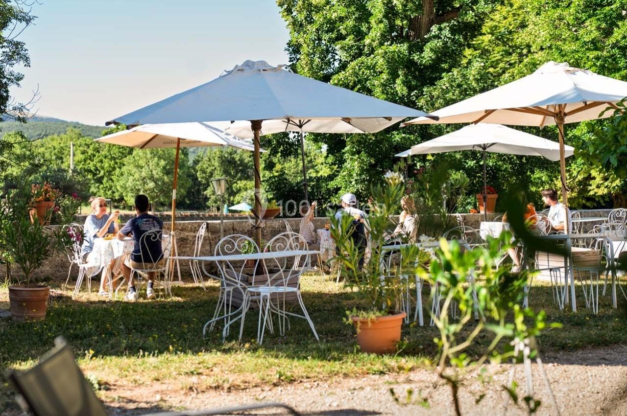 Terrasse extérieure avec tables et chaises en fer forgé, ombragée par des parasols, entourée de verdure.