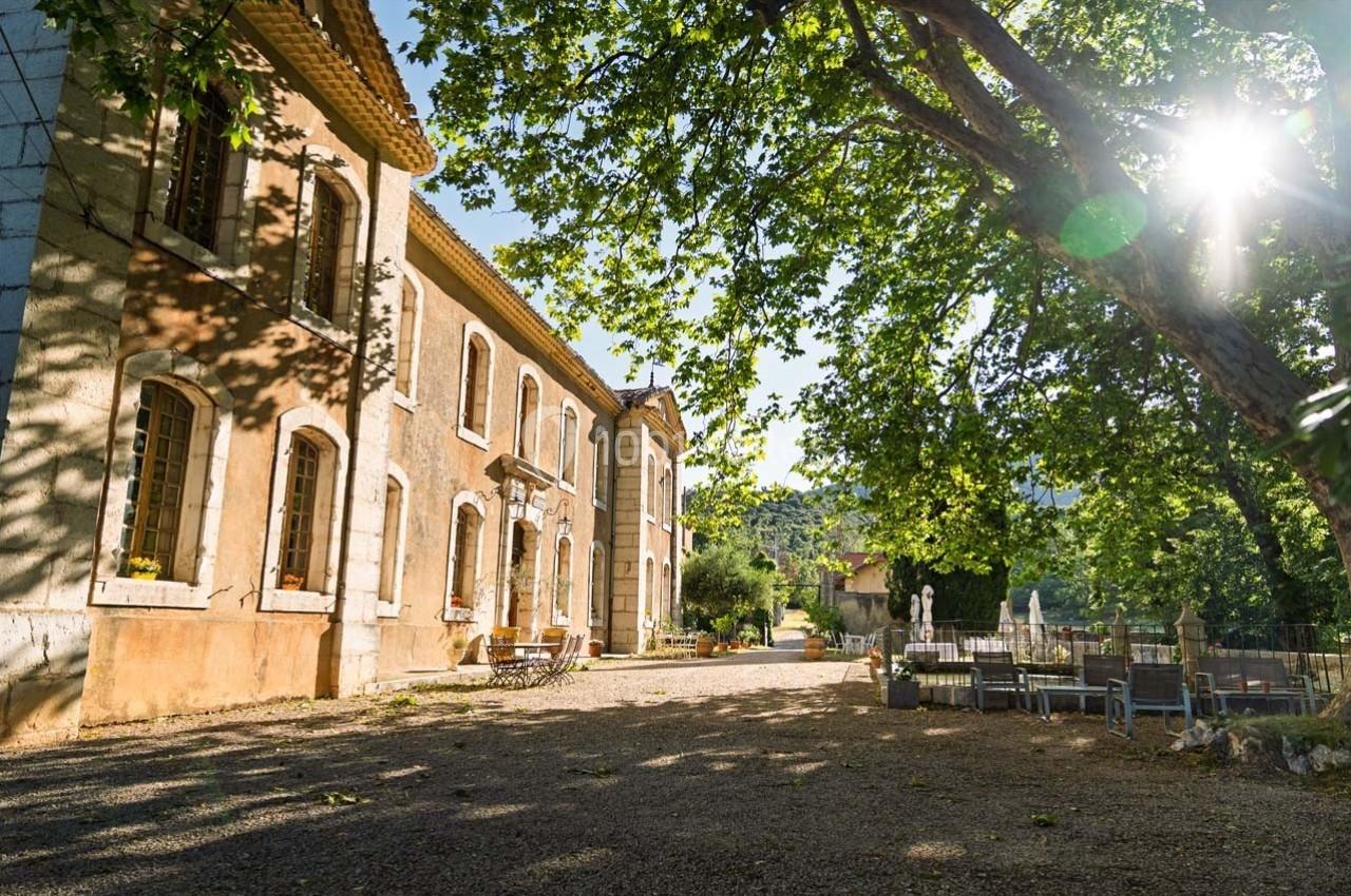 Bâtiment ancien en pierre entouré d'arbres, avec une cour ombragée et des tables en extérieur sous un soleil éclatant.
