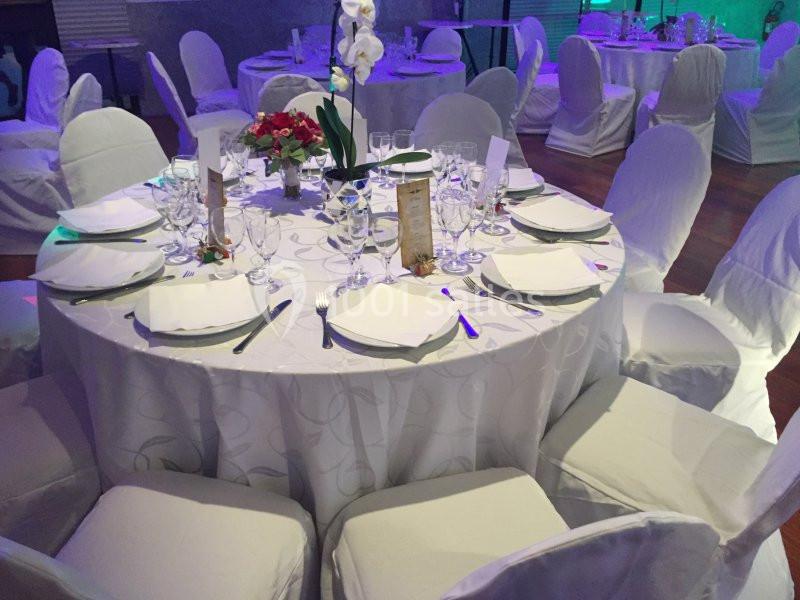 Table ronde dressée pour un événement, avec nappes blanches, chaises couvertes, vaisselle et décoration florale.
