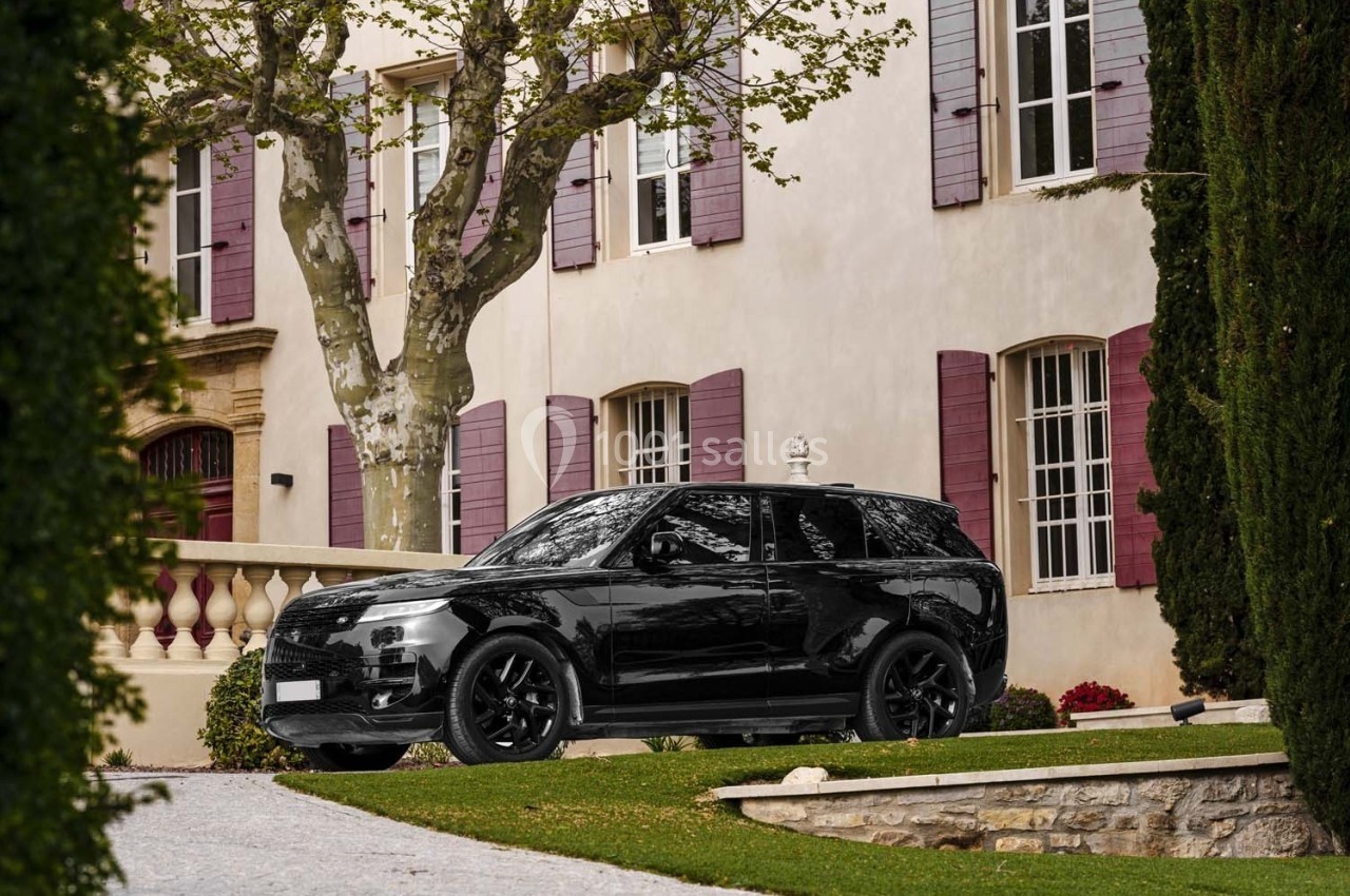 Voiture noire garée devant une maison beige aux volets pourpres, entourée d'arbres et de verdure.