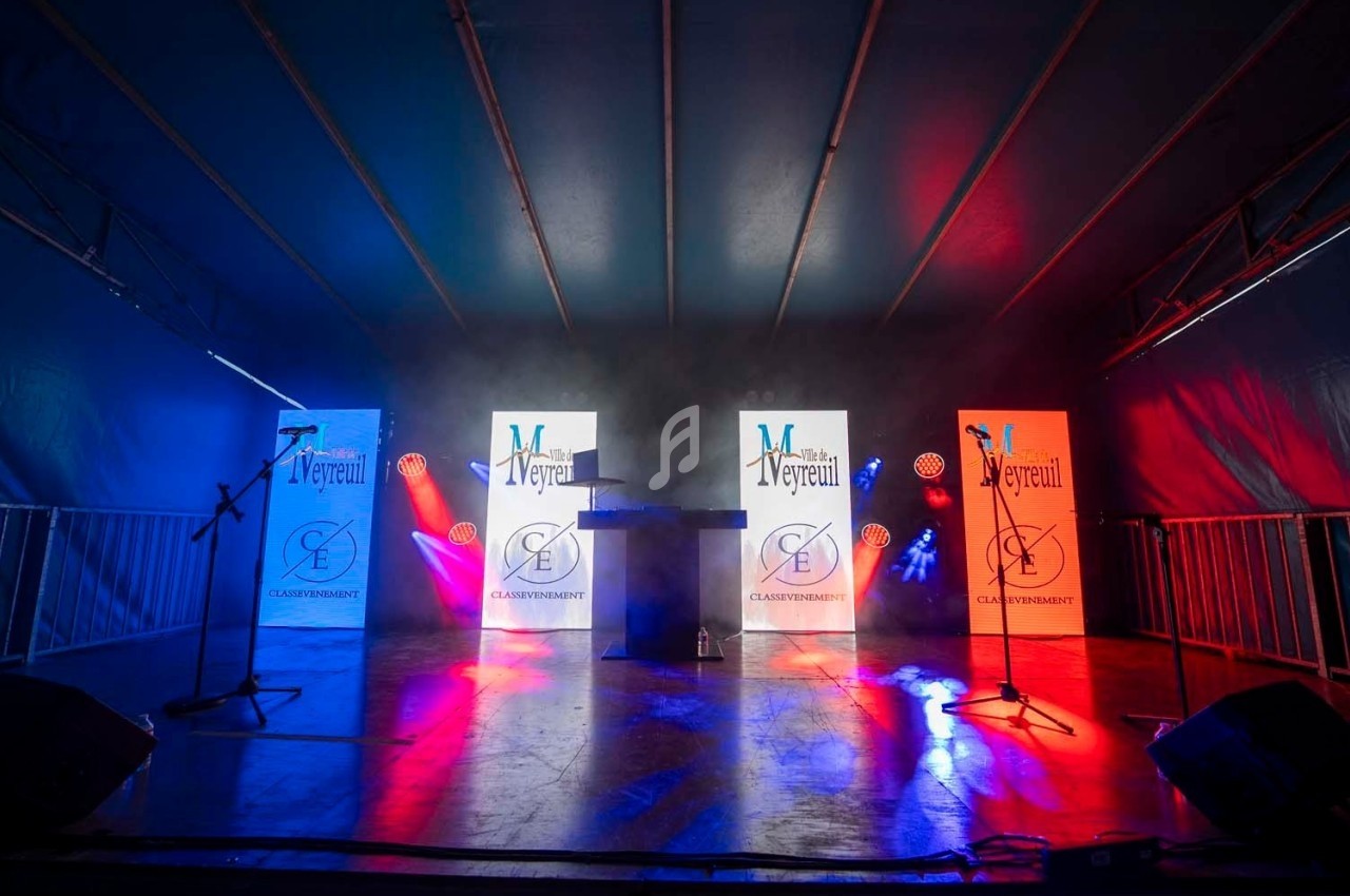 Scène éclairée en bleu, blanc et rouge avec deux micros, un pupitre central et des panneaux lumineux en arrière-plan.