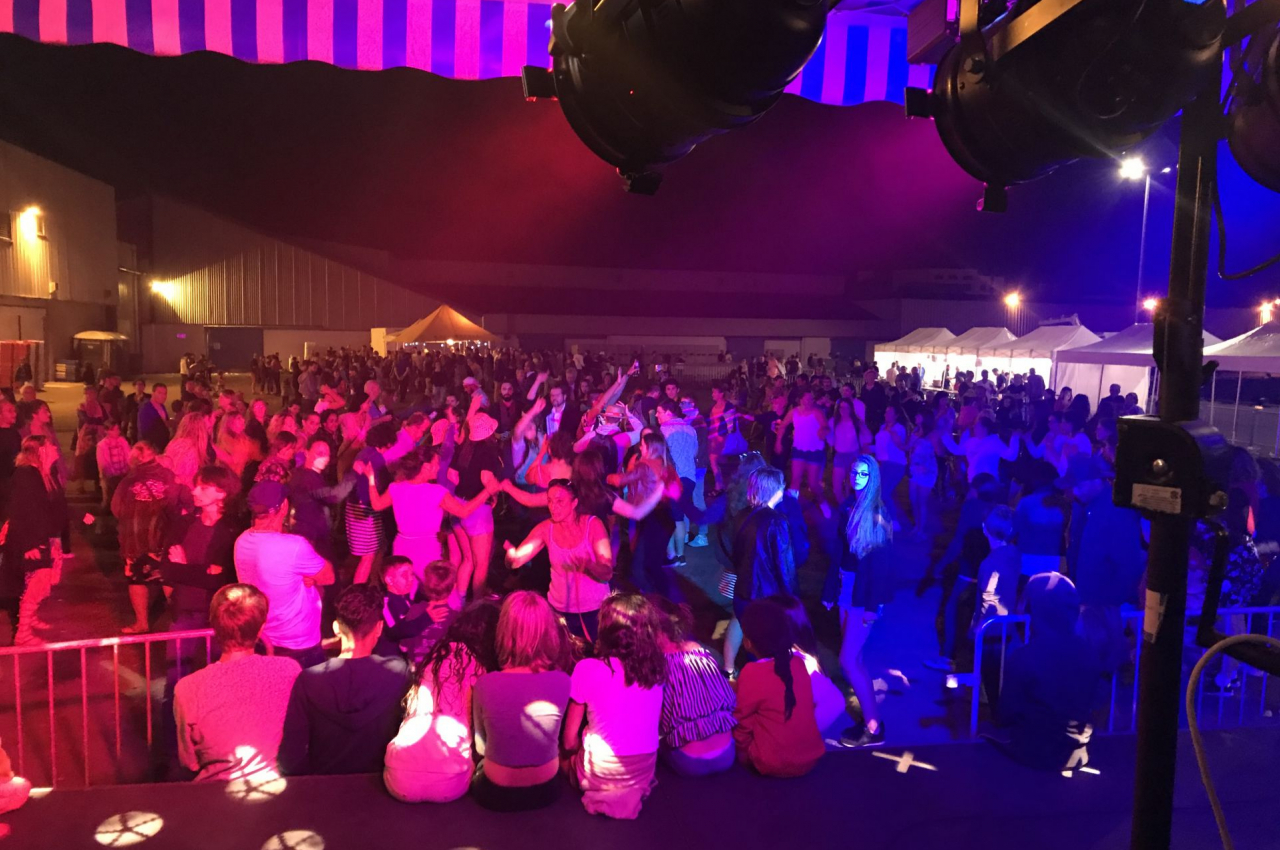 Foule dansant sous des éclairages colorés lors d'un événement nocturne en plein air avec des tentes en arrière-plan.