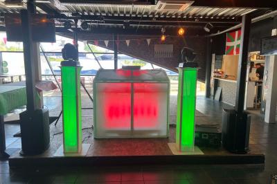 Stand de DJ avec éclairage LED rouge et vert, installé dans une salle avec décoration intérieure et lumière naturelle.