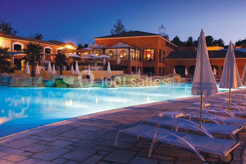 Piscine éclairée en soirée avec transats alignés au premier plan et bâtiments en arrière-plan.