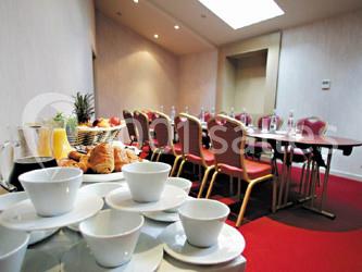 Salle de réunion avec table dressée, chaises rouges et buffet comprenant boissons, fruits et viennoiseries.