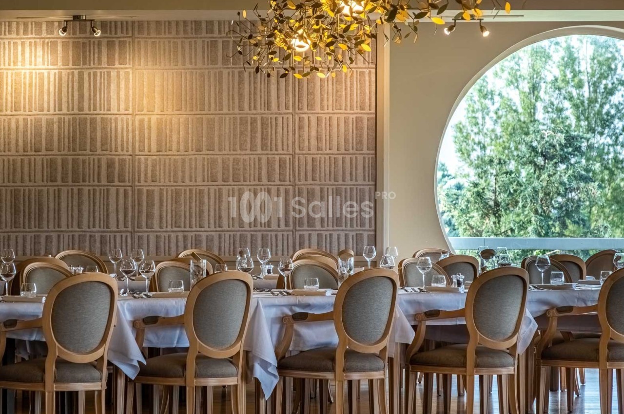 Salle de restaurant élégante avec de longues tables dressées, chaises en bois et grande fenêtre ronde donnant sur des arbres.