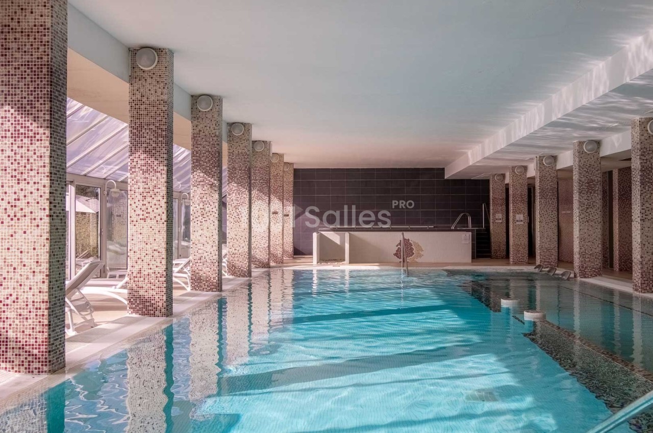 Piscine intérieure lumineuse entourée de colonnes carrelées, avec transats alignés près des baies vitrées.