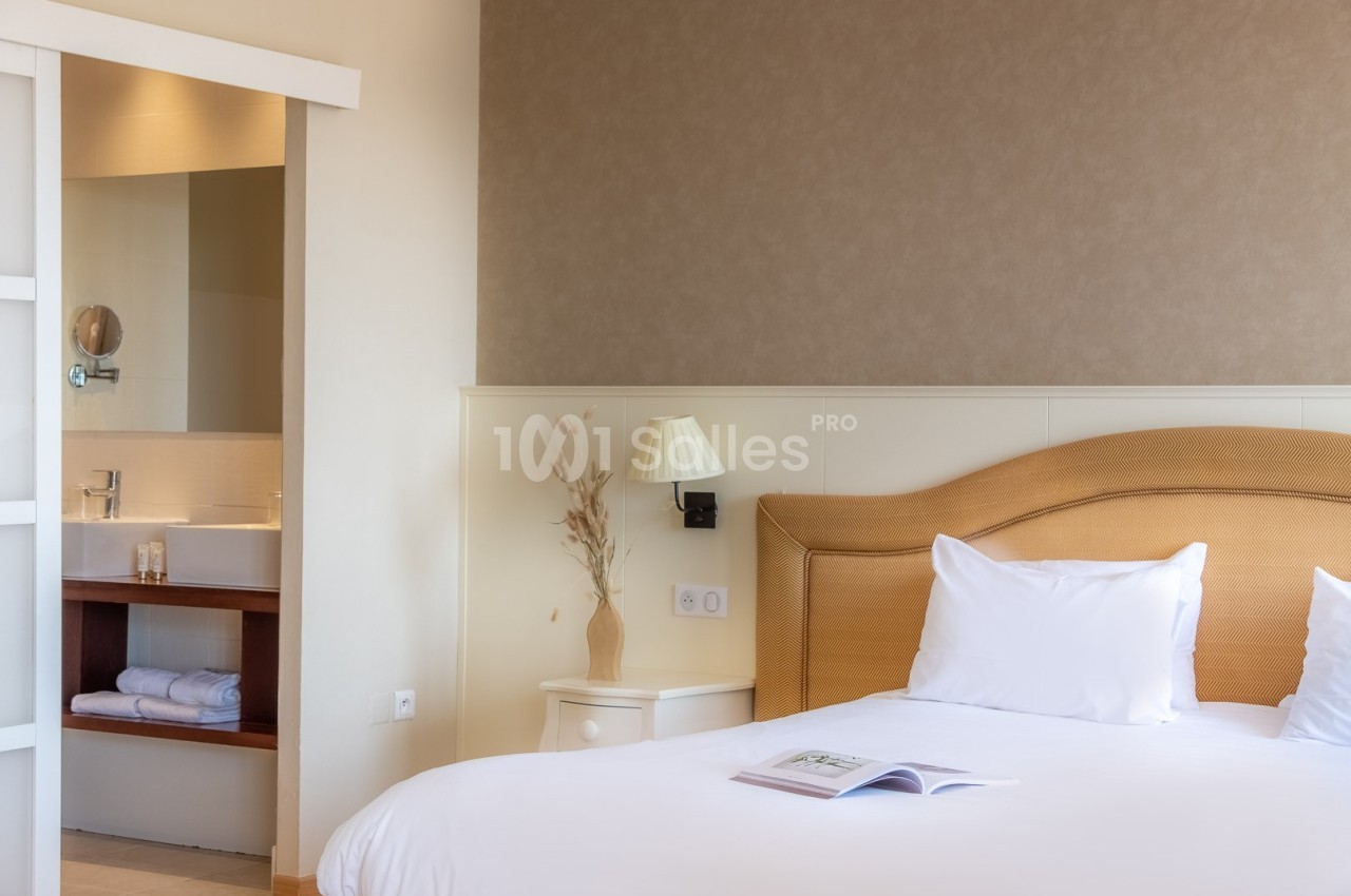Chambre lumineuse avec un lit double, une table de chevet, une lampe et une salle de bain attenante visible en arrière-plan.