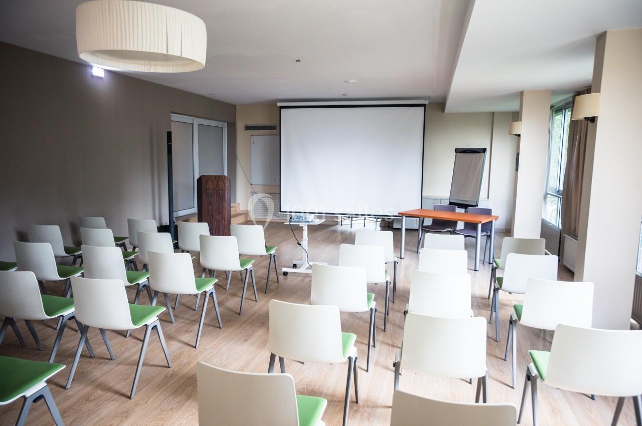 Salle de conférence lumineuse avec des rangées de chaises blanches, un écran de projection et un tableau de présentation.