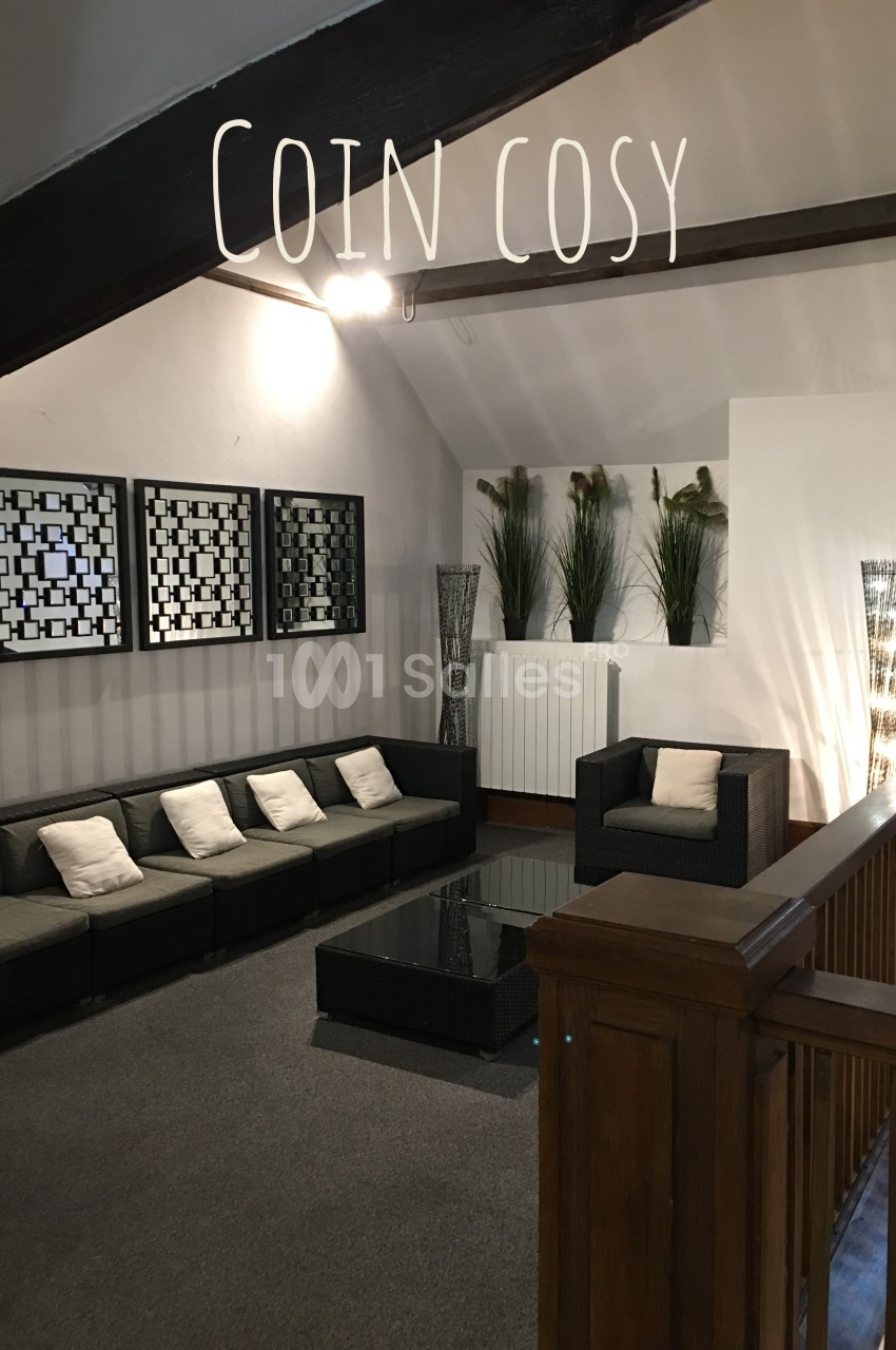 Espace salon avec canapé et fauteuil en rotin, table basse en verre, décoré de plantes et cadres muraux.