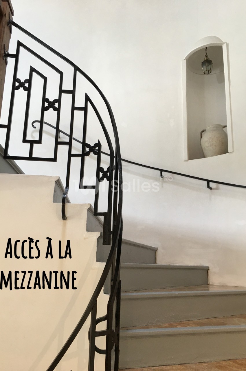 Escalier en colimaçon avec rampe en fer forgé menant à une mezzanine, mur blanc et niche décorative.