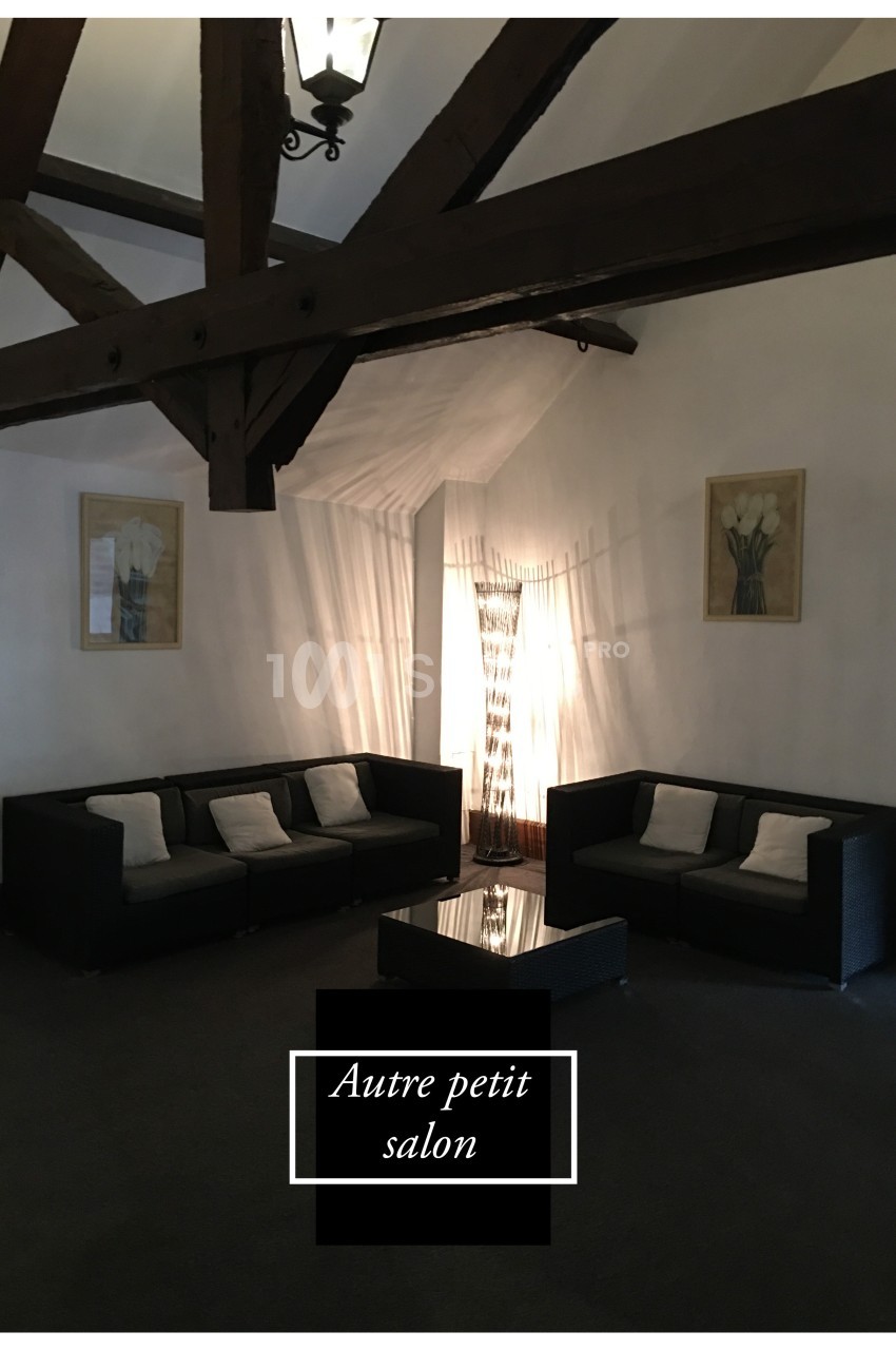 Deux canapés noirs avec coussins entourent une table basse dans un salon à poutres apparentes, éclairé par une lampe.